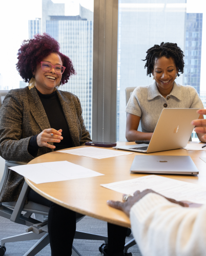 Colleagues laughing and engaged in a meeting around a conference table with laptops and documents.