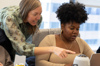 An On-Ramper in a grey, button-up shirt looks at a laptop perched on his lap as an On-Ramper in a plaid shirt looks over his shoulder. 