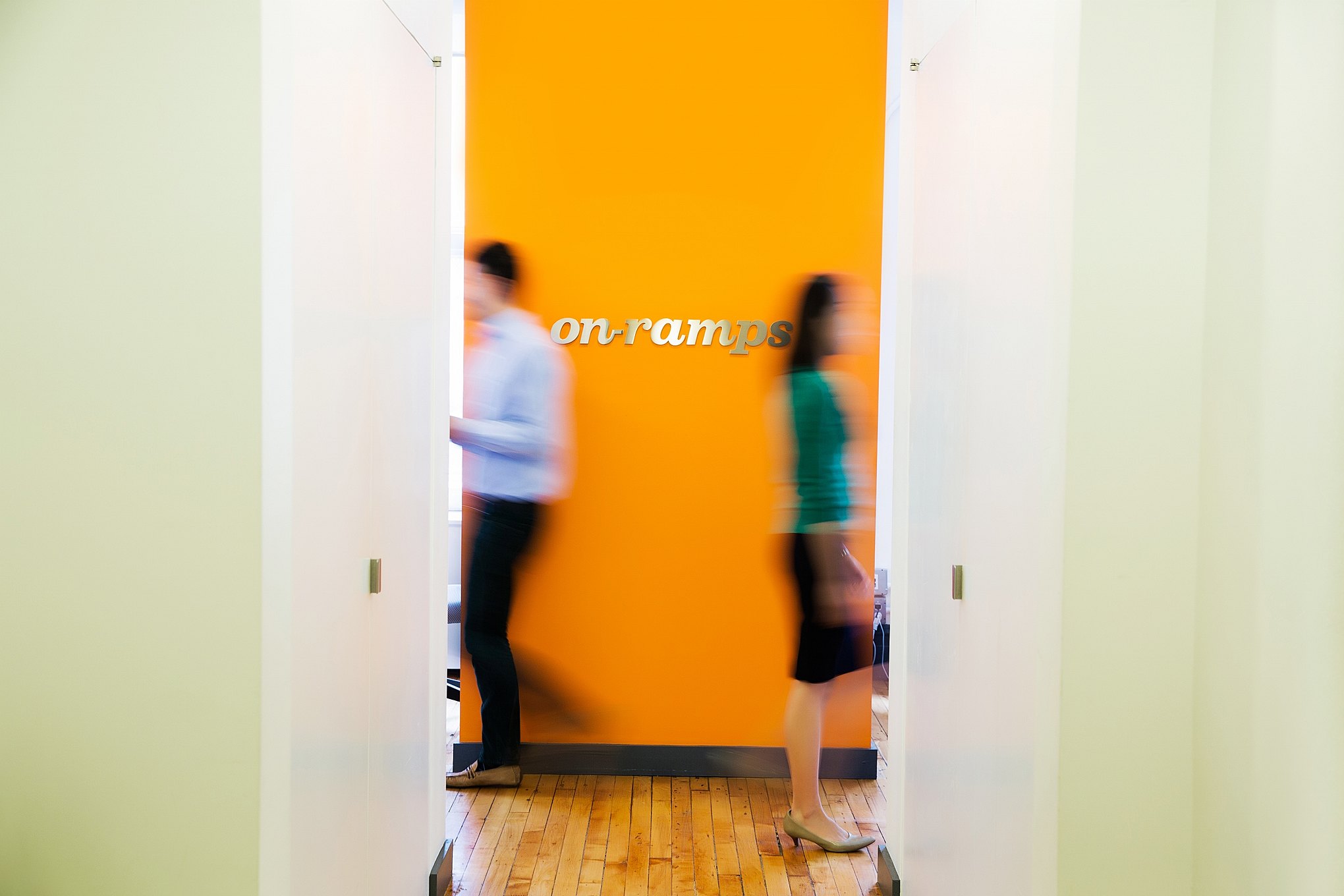People walking past the On-Ramps logo hanging on an orange  wall in the New York City office